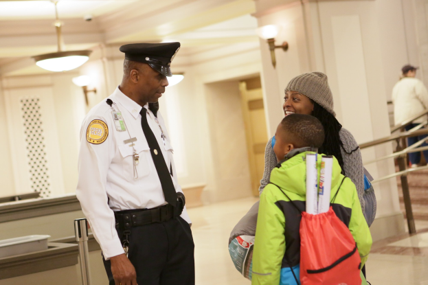 Ein Polizeibeamter spricht mit einer Frau mit Mütze und einem Kind an einem Flughafen, mit anderen Personen im Hintergrund bei beleuchteten Wandarmaturen und Treppen.