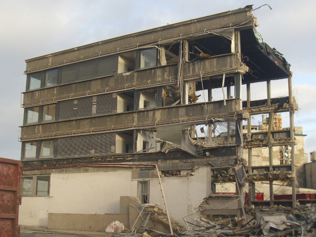 Ein teilweise abgerissener Bau inmitten einer Straße mit einem Container auf der linken Seite, zerbrochene Fenster und herumliegende Trümmer, unter einem bewölkten Himmel.