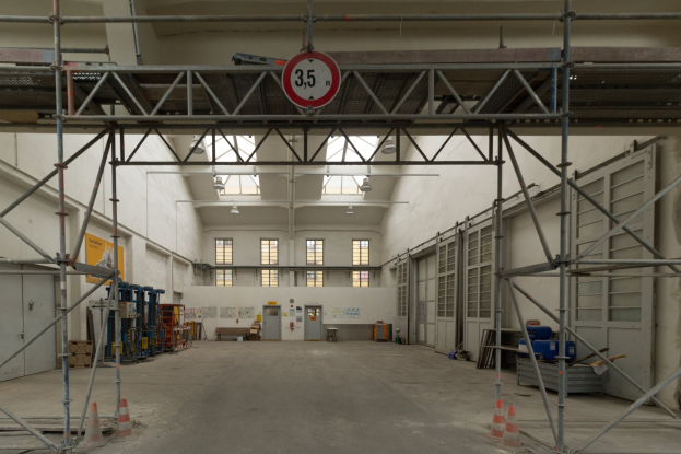 Innenansicht eines großen Lagerhauses mit ausgedehnten Gerüsten, Verkehrskegeln, Metallstangen, einem Schild, Maschinen, einer Tür, Fenstern, Wänden, Deckenleuchten und Dach.