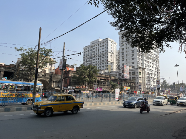 Gelbes Taxi auf einer belebten Straße in Mumbai mit hohen Gebäuden, Bäumen und Strommasten im Hintergrund.