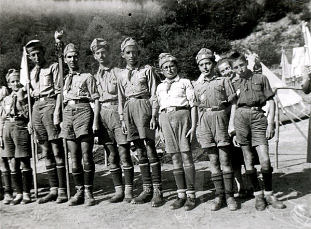Schwarz-weiß-Foto einer Gruppe von Pfadfindern in Uniform, die Fahnen halten, mit Zelten und Bäumen im Hintergrund.