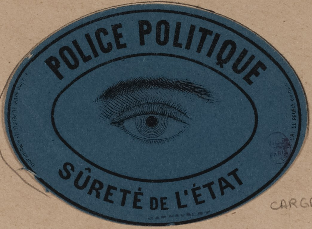 Brauner Karton mit einem blauen Aufkleber mit der Aufschrift "Polizei Politique Surete de L'État" und einem mittigen Auge-Symbol.
