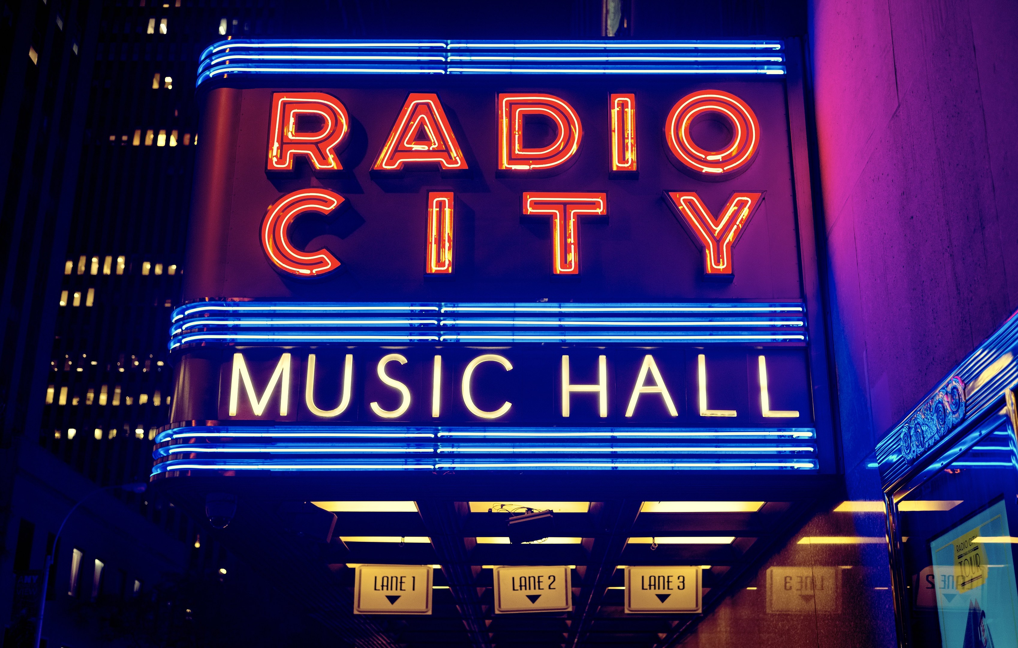 Außensicht des Radio City Music Hall in New York City bei Nacht, hell erleuchtet und von hohen Gebäuden flankiert, mit einem "Radio City"-Schild und zusätzlichen Schildern auf der rechten Seite.