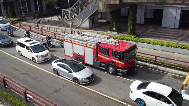 Eine Feuerwehrauto fährt auf einer Straße mit Autos, Motorrädern und anderen Fahrzeugen neben einer Begrenzung, mit Gebäuden, Bäumen und verschiedenen städtischen Elementen im Hintergrund.