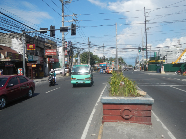 Eine belebte Stadtstraße mit Autos, Bussen, Motorrädern, Strommasten, Ampeln, Gebäuden mit Schildern, Bäumen und einem bewölkten Himmel.