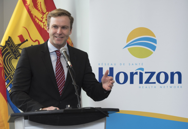 Ein Mann mit einem schwarzen Blazer, weißem Hemd und roter Krawatte steht an einem Podium mit einem Mikrofon und einem Banner im Hintergrund.