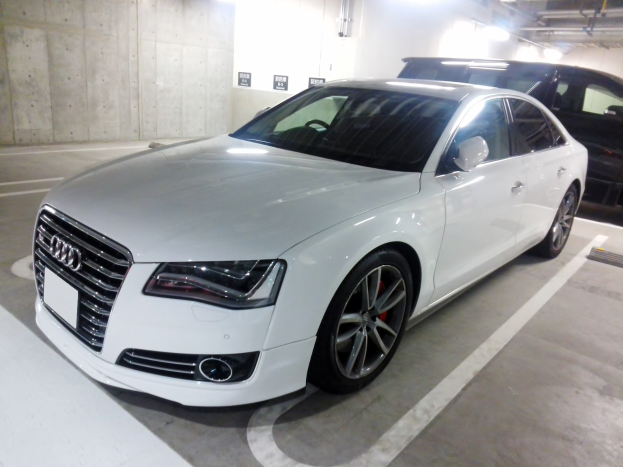 Ein weißer Audi S8 in einer Parkgarage neben einem schwarzen Auto geparkt, mit Garagenwänden, Deckenrohren und Leuchten im Hintergrund.