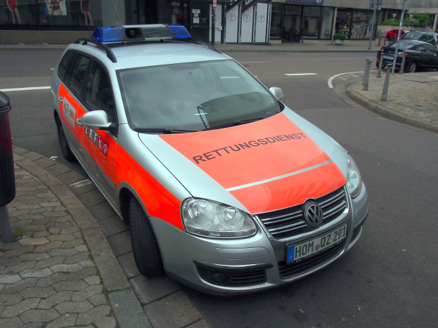 Ein Polizeiwagen am Straßenrand geparkt mit einem Müllcontainer auf dem Gehweg links und Gebäuden, Pfählen, Plakaten, Pflanzen, Bäumen und anderen Objekten im Hintergrund.
