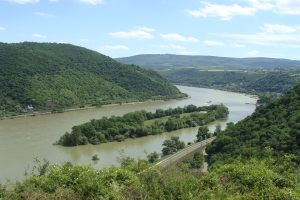 Ein malerischer Blick auf den Rhein, der sich durch eine grüne Landschaft schlängelt, mit einer Straße daneben und einem Himmel voller weißer, flauschiger Wolken.