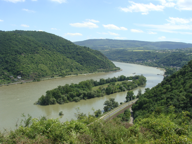 Ein malerischer Blick auf den Rhein, der sich durch eine grüne Landschaft schlängelt, mit einer Straße daneben und einem Himmel voller weißer, flauschiger Wolken.