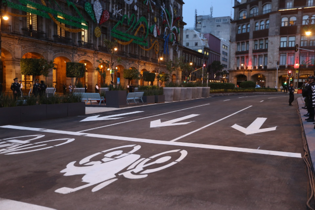 Stadtstraße mit einem ausgewiesenen Fahrradweg, umgeben von Gebäuden, Straßenmöbeln, Fußgängern auf dem Gehweg, Grünflächen und einem klaren blauen Himmel.