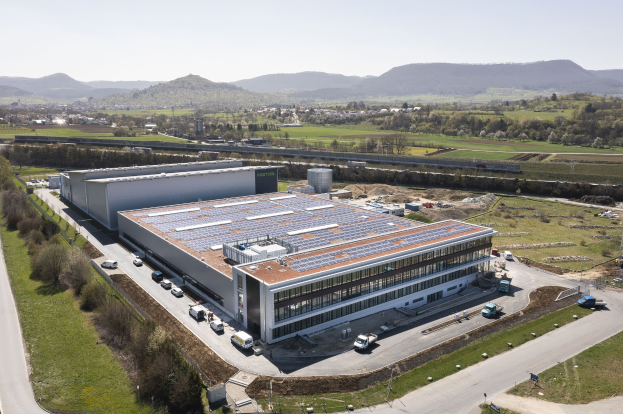 Luftbild einer großen Halle mit Solarmodulen auf dem Dach, umgeben von Fahrzeugen, Bäumen, Gras und Strommasten, mit Hügeln und einem klaren blauen Himmel im Hintergrund.