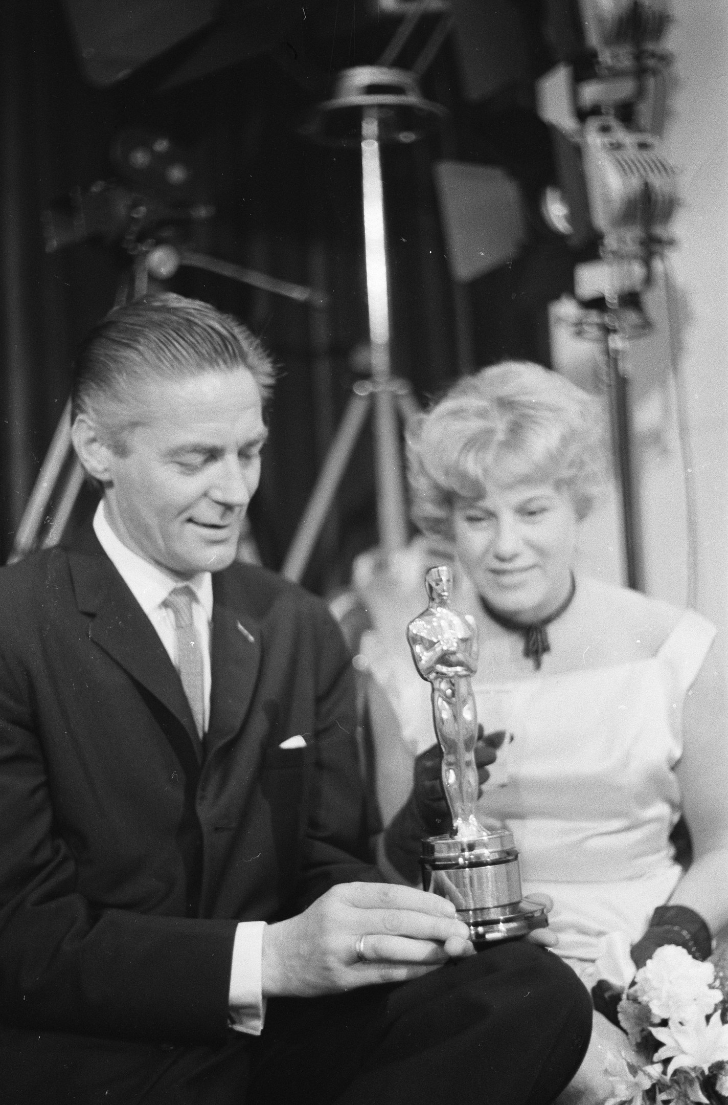 Ein Mann in einem schwarzen Blazer, einem weißen Hemd und einer Krawatte hölt eine Oscar-Statue, waehrend er neben einer Frau in formeller Kleidung sitzt, die einen Blumenstrauß hölt, mit Hintergrundbeleuchtung in einem Schwarz-Weiß-Bild.