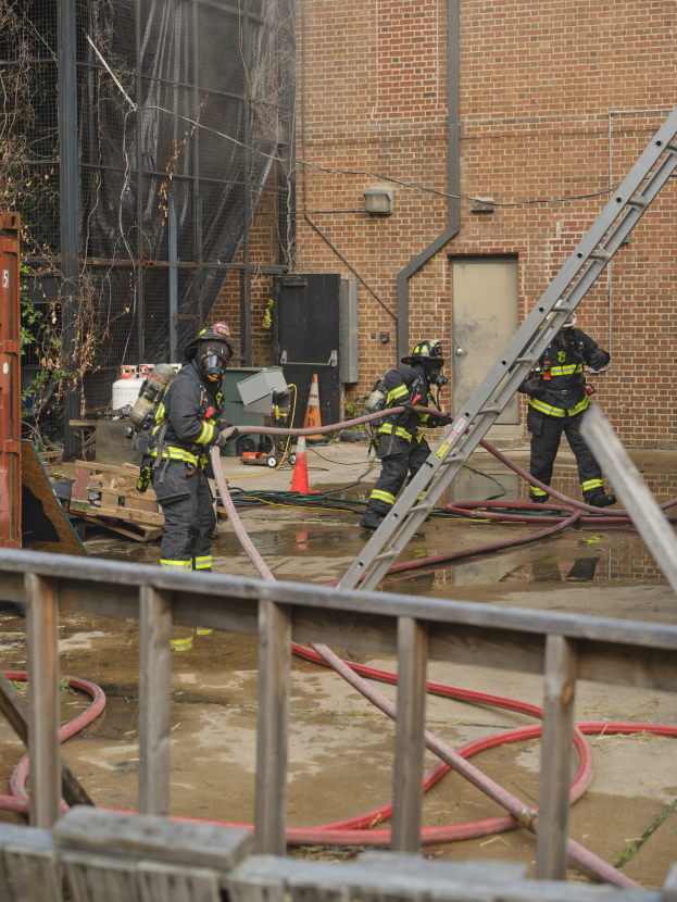 Feuerwehrleute mit Helmen arbeiten daran, ein Gebäudefeuer zu löschen, während sie Schläuche halten, mit einem Metallzaun, verstreuten Rohren, einem Container, einem Verkehrskegel, Gegenständen, Fenstern, einer Tür, einem Metallrahmen, Drähten, einem Baum und dem Himmel im Hintergrund.