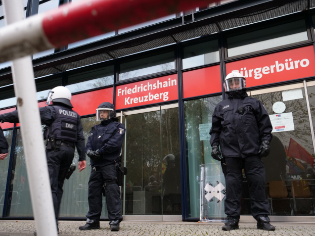 Polizeibeamte in Helmen und Handschuhen vor einem Gebäude mit Glaswänden und -türen, neben einem Pfahl, mit "Friedrichshain Kreuzberg"-Text auf Schildern im Hintergrund.