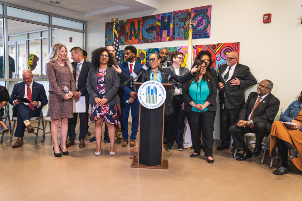 Gruppe von Menschen in einem Raum mit einem Podium, Mikrofon und einer Tafel mit der Aufschrift "Sancho Stadtverordneter tritt dem Stadtrat bei", umgeben von Flaggen, Sitzplätzen und einem Feuerüextinguisher.