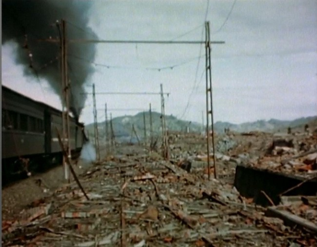 Ein Zug fährt durch ein beschädigten Gebiet mit Rauch, der aus ihm aufsteigt, umgeben von Masten und Drähten, gegen Hügel unter einem klaren Himmel, mit verstreuten Trümmern am Boden.