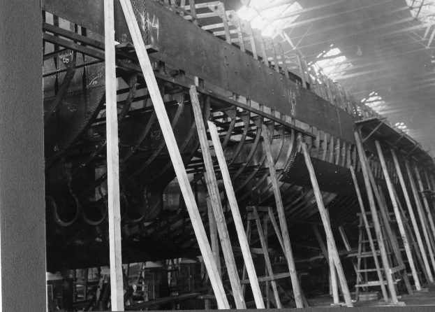 Schwarzes und weißes Foto eines Boots in der Herstellung in einer Fabrik, mit Holzpfählen, Leitern und anderen Gegenständen, die auf dem Boden unter einem sichtbaren Dach verstreut sind.