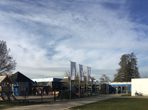 Großes Gebäude mit einem grasbewachsenen Bereich davor, umgeben von einem Metallzaun, mit einem Weg, Bäumen, Pfosten, Bannern, einem geparkten Fahrrad, nahegelegenen Häusern und einem bewölkten Himmel, als das neue Spielgelände der Schule identifiziert.