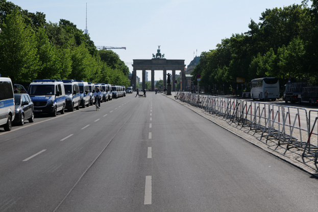 Lange Reihe von Polizeiwagen, die entlang einer Straße vor dem Brandenburger Tor geparkt sind, mit Fahrradfahrern und Fußgängern in der Nähe, Absperrungen, Bäumen und dem Torbogen und den Statuen im Hintergrund.