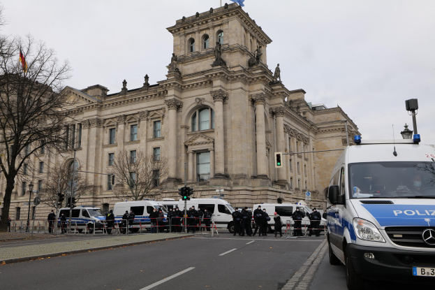 Eine Gruppe von Polizisten steht vor dem Reichstaggebäude in Berlin, Deutschland, mit Fahrzeugen, einem Zaun, Verkehrsampeln, Laternenpfählen, Bäumen und Fahnen im Hintergrund, unter einem klaren Himmel.