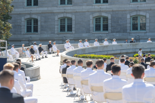 Gruppe von Menschen auf Stühlen vor einem Gebäude mit Fenstern, umgeben von Grün, während einer Zeremonie an der US Naval Academy mit einem Podium und Mikrofon im Vordergrund.