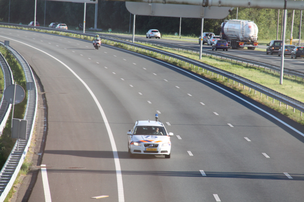 Ein Auto auf einer Autobahn mit einem stehenden Polizeiauto am Straßenrand, andere Fahrzeuge und Bäume, Geländer, Pfosten und Bretter im Hintergrund.