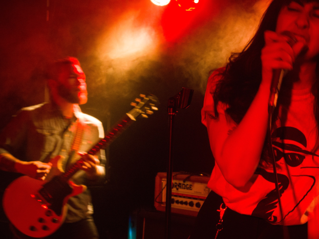 Eine Frau singt in ein Mikrofon auf der Bühne, während ein Mann im Hintergrund eine akustische Gitarre spielt und Deckenlampen die Szene beleuchten.