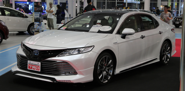Weißer Toyota Camry auf einer Autauschau präsentiert, mit Menschen in bunten Kleidern im Vordergrund und Schildern, Lichtern und Glastüren im Hintergrund.
