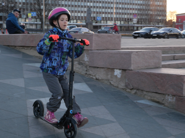 Ein junger Junge in Helm und Handschuhen fährt ein Scooter auf einem Gehweg mit Stufen, Fahrzeugen, Menschen, Bäumen, Pfählen, Brettern, Gebäuden und einem klaren blauen Himmel im Hintergrund.