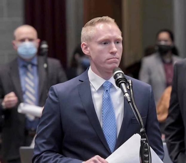 Ein Mann in Anzug und Krawatte spricht an einem Rednerpult mit Papieren in der Hand, während einige Menschen hinter ihm stehen, vor einer Vorhangwand.