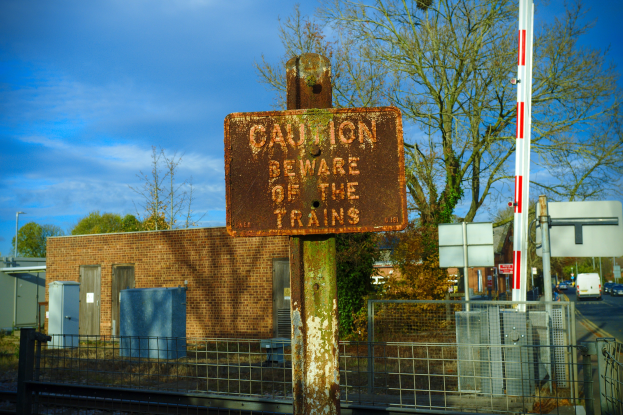 Vorsichtsschild an einem Bahnübergang mit Bäumen, Strommasten, einem Gebäude, Containern, Fahrzeugen auf einer Straße und einer bewölkten Himmel.