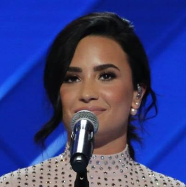 Demi Lovato steht mit einem Mikrofon auf der Bühne bei den Grammys, trägt ein schwarzes Kleid und einen sleeken Bob, mit einem blauen Hintergrund dahinter.