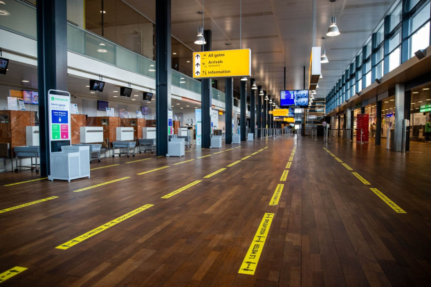 Ein langer Flughafenkorridor mit gelbem Absperrband auf dem Boden, Säulen, Informationsanzeigen, Bildschirme, Lampen, Glaswände und Fenster, mit einer Person im Hintergrund.