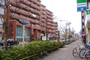 Eine Stadtstraßenszene mit parkenden Fahrrädern, Gebäuden, Straßenlaternen, Fahrzeugen, Passanten, Grünflächen und einem klaren blauen Himmel.