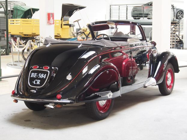 Schwarzer und roter Wagen in einer Garage mit anderen Fahrzeugen, Pfählen, Ketten und Gegenständen im Hintergrund, neben einem wandmontierten Feuerlöscher.