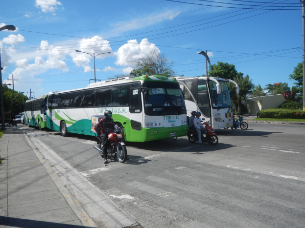 Ein grüner und weißer Shuttlebus parkt am Straßenrand mit ein paar Motorrädern davor, einem grasbewachsenen Fußweg links und Gebäuden, Bäumen, Laternenmasten und einem klaren blauen Himmel im Hintergrund.