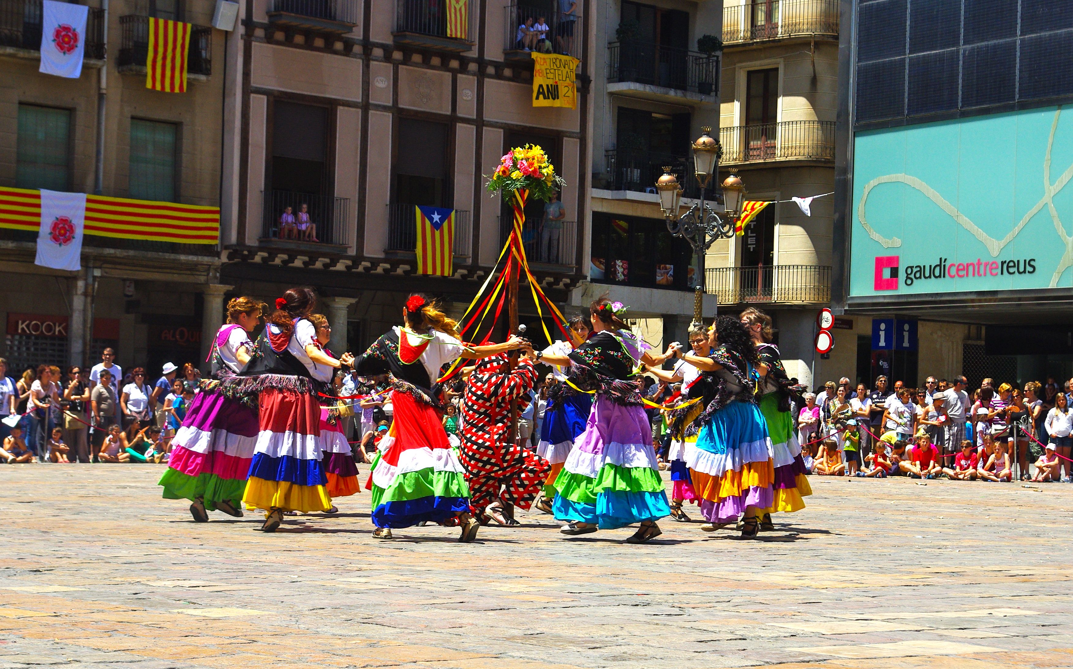 Eine Gruppe von Frauen in bunten Kleidern tanzt auf der Straße, umgeben von einer Menge Menschen, mit Gebäuden, Geländern, Balkonen, Fahnen, einem Laternenmast und einer Tafel mit Schrift im Hintergrund.