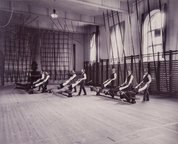 Schwarzes und weißes Foto von Menschen, die Pilates-Übungen in einem Fitnessstudio mit einer Wand, Fenstern auf der rechten Seite und Deckenleuchten im Hintergrund durchführen.