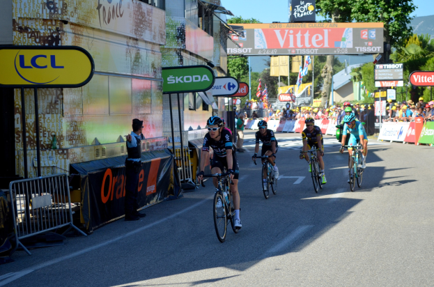 Gruppe von Radfahrern mit Helmen und Schutzbrillen, die eine Straße mit Absperrungen, Schildern und Tafeln mit Text entlangfahren, während eine Person links steht; Bäume, Gebäude, Flaggen und Pfosten sind im Hintergrund unter einem klaren blauen Himmel zu sehen.