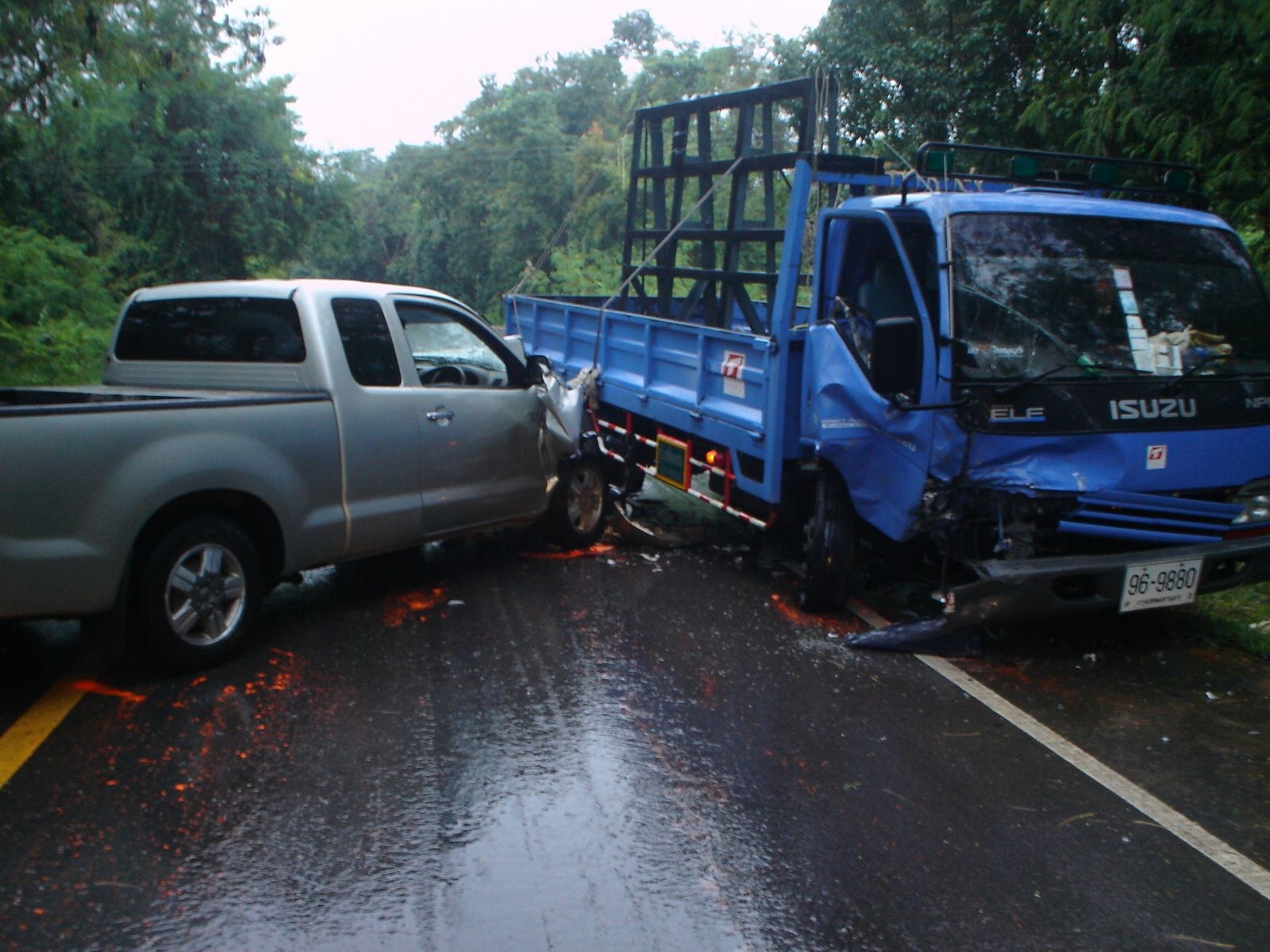 Ein schwerbeschädigtter Lkw mit eingedellter Front und verbeulter Karosserie liegt auf der Seite einer Straße, umgeben von Bäumen unter einem klaren blauen Himmel.