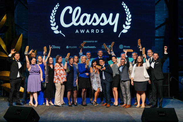 Eine Gruppe von Menschen auf einer Bühne, die lächeln und Auszeichnungen halten, mit Lautsprechern unten und einem Hintergrundbild, das "Classy Awards 2019" anzeigt.