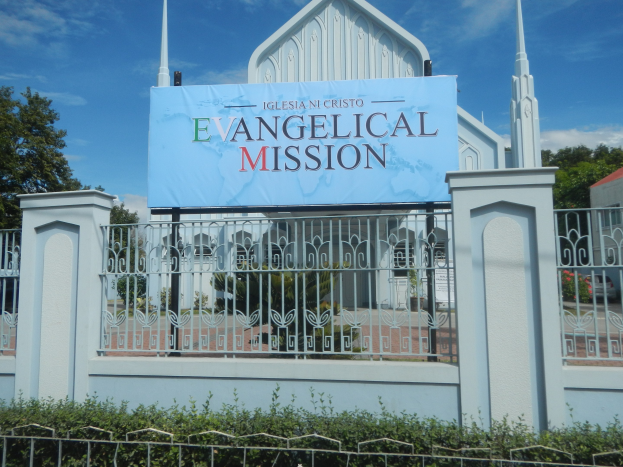 Kirche mit einem Metallzaun, einem Schild mit der Aufschrift "Evangelische Mission", umgeben von Bäumen, Pflanzen und einem benachbarten Haus unter einem bewölkten Himmel.