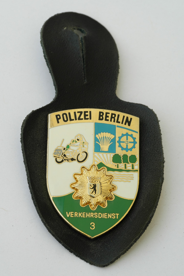 Ein schildförmiges Polizeibadge mit 'Polizei Berlin' in schwarzer fetter Schrift auf einem blauen Hintergrund mit einem weißen Stern und Rahmen, das auf einer Oberfläche liegt.