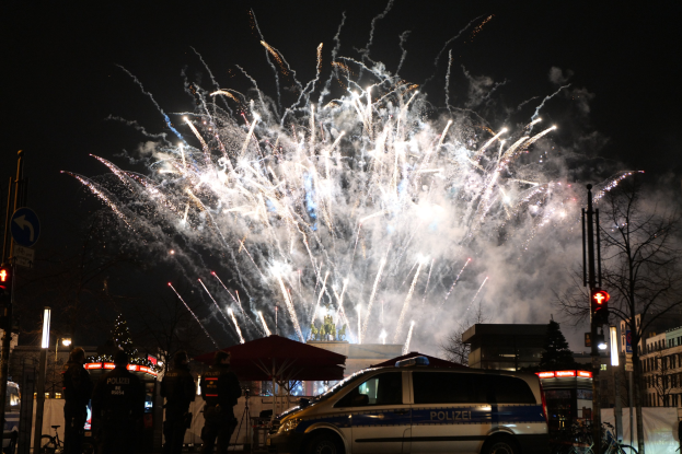 Polizeiauto vor einer Menschenmenge während einer Silvesterfeier in Berlin geparkt, mit farbigen Feuerwerkskörpern am Himmel.