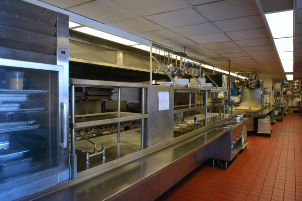 Gewerbeküche mit Edelstahl-Theke, Deckenbeleuchtung, hängenden Küchengeräten, Spüle, Wasserhähnen, Schränken und diversen Gegenständen in Regalen.