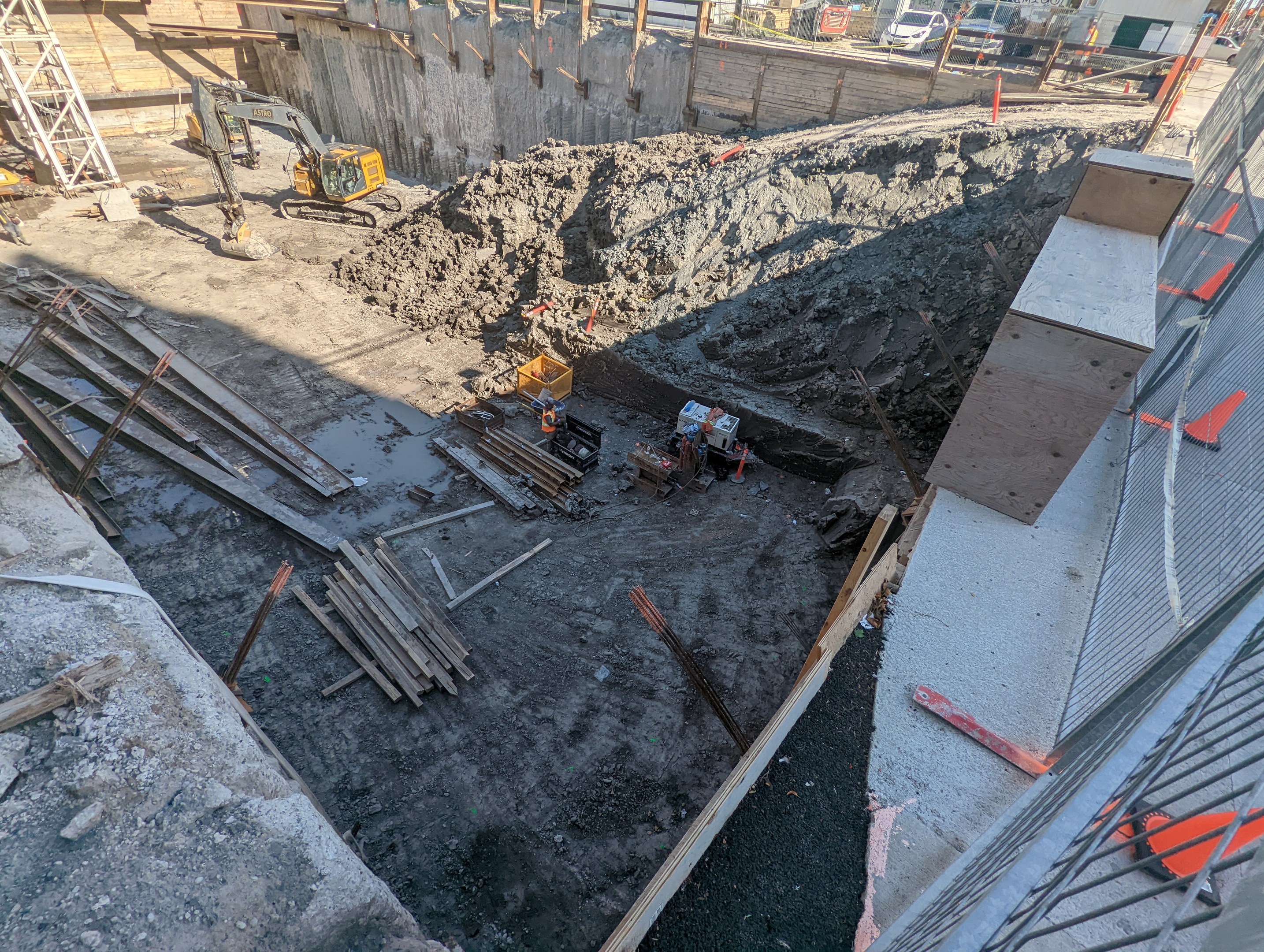 Baustelle mit einem großen Schutthaufen, umgeben von Kränen, Holzstöcken und anderem Equipment, umgeben von einem Maschendrahtzaun, mit Gebäuden, Fahrzeugen, Pfählen, Bäumen und Himmel im Hintergrund.