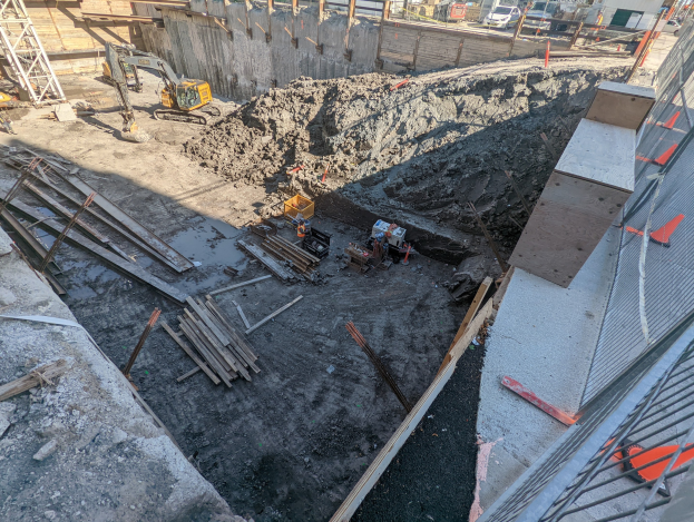 Baustelle mit einem großen Schutthaufen, umgeben von Kränen, Holzstöcken und anderem Equipment, umgeben von einem Maschendrahtzaun, mit Gebäuden, Fahrzeugen, Pfählen, Bäumen und Himmel im Hintergrund.