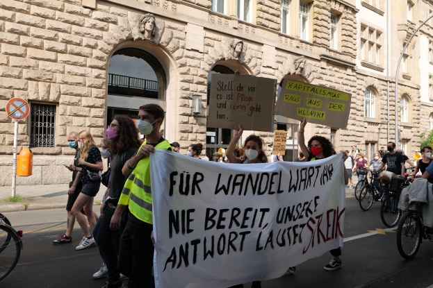 Eine Gruppe von Menschen marschiert bei einer Protestaktion gegen die Friedensbewegung in Berlin, einige halten Schilder und fahren Fahrräder, im Hintergrund ein Gebäude mit Bögen, Säulen und Skulpturen.