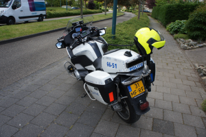 Ein Polizeimotorrad am Straßenrand geparkt mit einem Helm darauf, umgeben von Bäumen, Polen, Gebäuden, Pflanzen, Gras, Steinen und anderen Fahrzeugen.